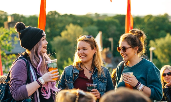 Three women smiling and chatting while holding drinks at an outdoor event, with greenery and flags in the background.